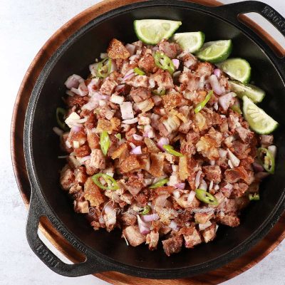 Crispy pork belly sisig with lime wedges in cast iron pan